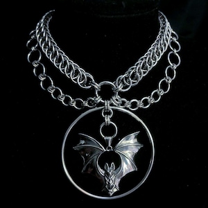 Könnte beinhalten: Eine silberfarbene Kettenhemd-Halskette mit einem mehrschichtigen Design aus ineinandergreifenden Metallringen. Ein großer kreisförmiger Anhänger hängt in der Mitte und enthält eine detaillierte Fledermausfigur aus Metall mit ausgebreiteten Flügeln.