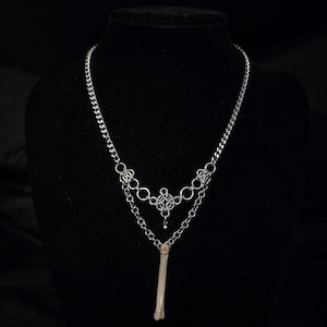 Op de afbeelding: Een zilverkleurige ketting met een gelaagd ontwerp. De bovenste laag heeft een ketting en decoratieve schakels, terwijl de onderste laag een bottenhanger heeft. De ketting wordt gepresenteerd tegen een zwarte achtergrond.