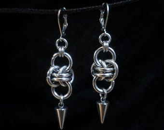 May include: A pair of silver-toned earrings. Each earring features a series of interconnected rings, culminating in a pointed, cone-shaped dangle. The earrings are suspended against a dark background, highlighting their metallic sheen.