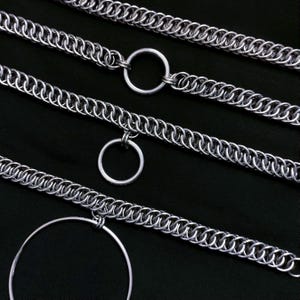 Könnte beinhalten: Nahaufnahme mehrerer silberner Kettenhemden-Halsketten mit kreisförmigen Anhängern. Die Ketten bestehen aus kleinen, ineinandergreifenden Metallringen, die ein strukturiertes, metallisches Aussehen erzeugen. Die Anhänger sind einfache, runde Reifen.