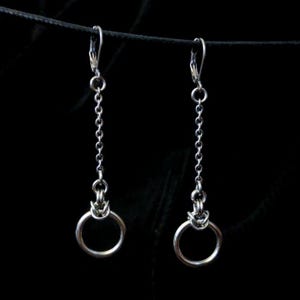 Könnte beinhalten: Ein Paar silberfarbene Hängeohrringe. Jeder Ohrring hat einen kleinen Bügelverschluss, eine Kette und einen kreisförmigen Ring am unteren Ende. Die Ohrringe hängen vor einem schwarzen Hintergrund.