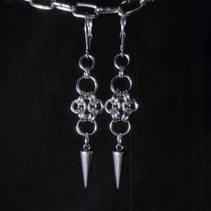 Könnte beinhalten: Ein Paar silberne Ohrringe mit einem Kettenglied-Design und einer spitzen Spitze. Die Ohrringe hängen an einer silbernen Kette.