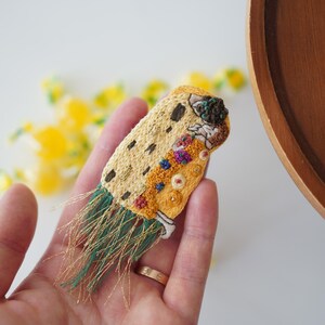 Puede incluir: Broche bordado con un dise&ntilde;o en hilos amarillos, marrones y verdes. El broche tiene forma rectangular con un patr&oacute;n detallado y flecos. El dise&ntilde;o incluye varios colores y texturas, con un anillo dorado visible.