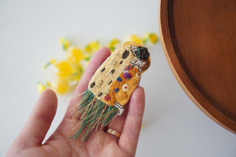Puede incluir: Broche bordado con un dise&ntilde;o en hilos amarillos, marrones y verdes. El broche tiene forma rectangular con un patr&oacute;n detallado y flecos. El dise&ntilde;o incluye varios colores y texturas, con un anillo dorado visible.