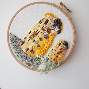 Puede incluir: Arte de bordado cosido a mano en un aro de madera. El dise&ntilde;o presenta una representaci&oacute;n estilizada de una pareja abraz&aacute;ndose, con tonos dorados, amarillos y marrones. La obra de arte est&aacute; adornada con cuentas e hilo de colores, sobre un fondo blanco.