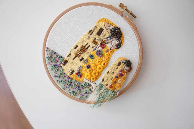 Puede incluir: Arte de bordado cosido a mano en un aro de madera. El dise&ntilde;o presenta una representaci&oacute;n estilizada de una pareja abraz&aacute;ndose, con tonos dorados, amarillos y marrones. La obra de arte est&aacute; adornada con cuentas e hilo de colores, sobre un fondo blanco.
