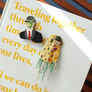 Puede incluir: Broches bordados sobre un libro. Uno representa una figura con traje y bomb&iacute;n, el otro un dise&ntilde;o abstracto y colorido con flecos. El libro tiene texto amarillo que dice "Viajando juntos a trav&eacute;s del tiempo cada d&iacute;a de nuestras vidas."