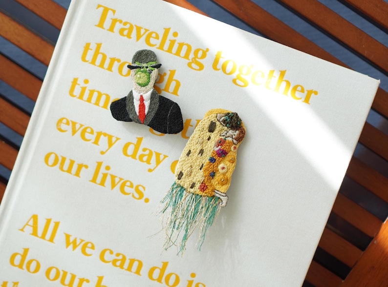 Puede incluir: Broches bordados sobre un libro. Uno representa una figura con traje y bomb&iacute;n, el otro un dise&ntilde;o abstracto y colorido con flecos. El libro tiene texto amarillo que dice "Viajando juntos a trav&eacute;s del tiempo cada d&iacute;a de nuestras vidas."