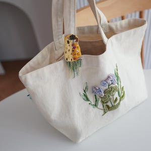 Puede incluir: Bolso tote color crema con dise&ntilde;o floral bordado en azul, verde y morado. Un peque&ntilde;o adorno decorativo cuelga de la abertura del bolso. El bolso tiene dos asas y est&aacute; sobre una superficie blanca.