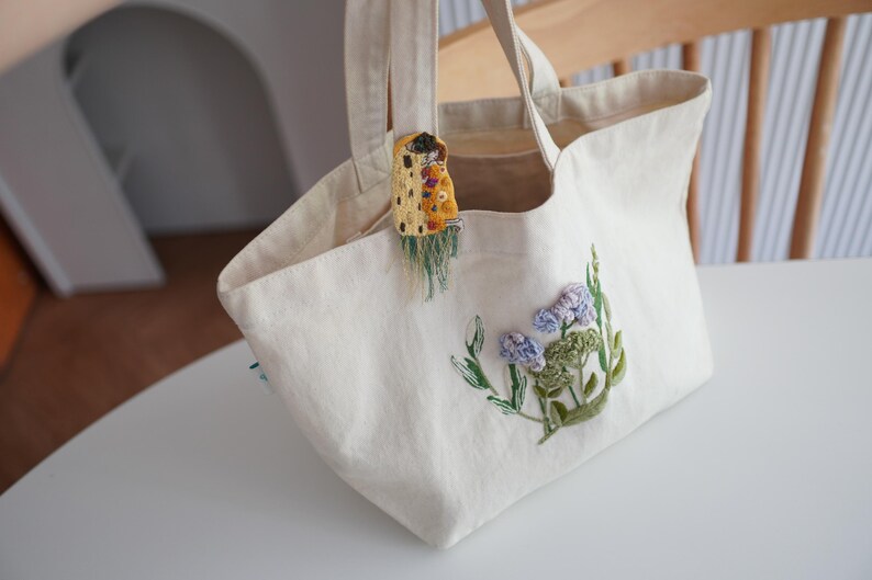 Puede incluir: Bolso tote color crema con dise&ntilde;o floral bordado en azul, verde y morado. Un peque&ntilde;o adorno decorativo cuelga de la abertura del bolso. El bolso tiene dos asas y est&aacute; sobre una superficie blanca.