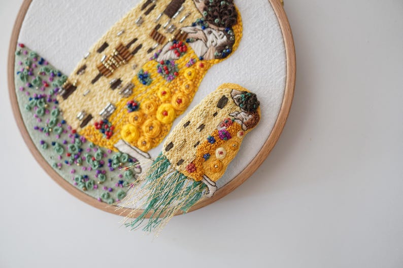 Puede incluir: Obra de arte bordada en un aro de madera. El dise&ntilde;o presenta una porci&oacute;n de "El Beso" de Gustav Klimt, con hilo dorado, amarillo y marr&oacute;n. La obra incluye cuentas y detalles texturizados, con un &aacute;rea de cuentas verdes y moradas.
