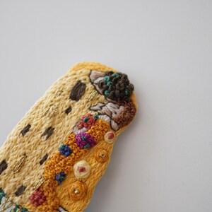 Puede incluir: Primer plano de una pieza de arte textil bordada a mano. La pieza presenta una base amarilla con detalles marrones y dorados, y bordados florales coloridos. Flecos verdes y dorados cuelgan de la parte inferior.
