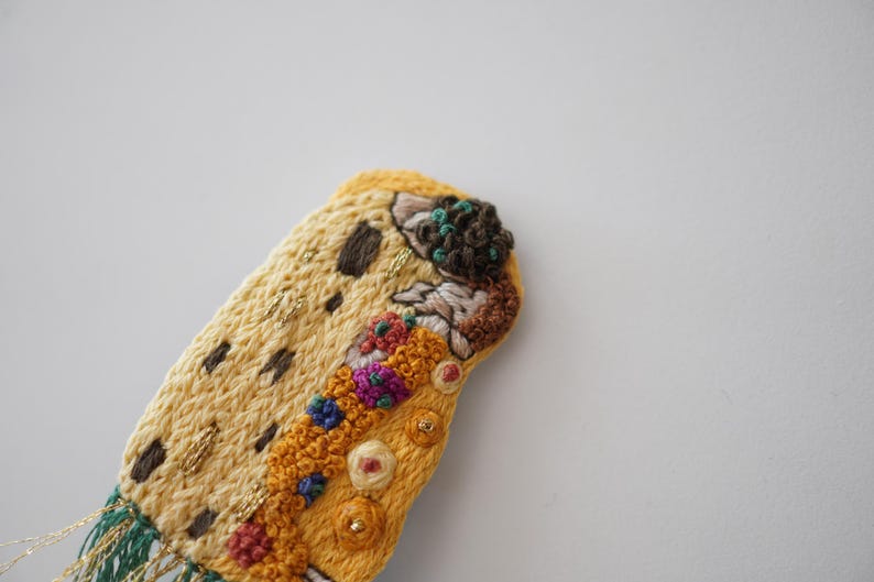 Puede incluir: Primer plano de una pieza de arte textil bordada a mano. La pieza presenta una base amarilla con detalles marrones y dorados, y bordados florales coloridos. Flecos verdes y dorados cuelgan de la parte inferior.