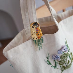 Puede incluir: Bolso tote de color crema con detalles florales bordados y un broche decorativo. El broche presenta un dise&ntilde;o amarillo y naranja con flecos verdes. El bolso tiene asas resistentes y est&aacute; hecho de una tela duradera.