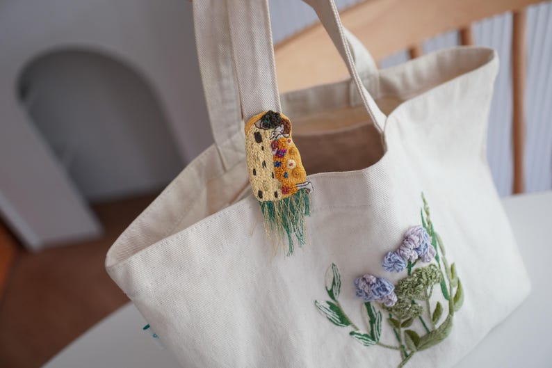 Puede incluir: Bolso tote de color crema con detalles florales bordados y un broche decorativo. El broche presenta un dise&ntilde;o amarillo y naranja con flecos verdes. El bolso tiene asas resistentes y est&aacute; hecho de una tela duradera.