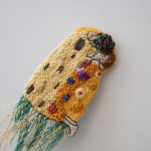 Puede incluir: Broche o elemento decorativo textil bordado. Presenta un dise&ntilde;o amarillo y beige con detalles en marr&oacute;n, rojo y azul. Cuenta con flecos de hilos verdes y dorados.