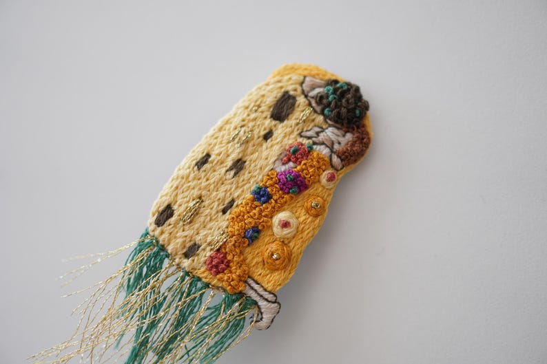 Puede incluir: Broche o elemento decorativo textil bordado. Presenta un dise&ntilde;o amarillo y beige con detalles en marr&oacute;n, rojo y azul. Cuenta con flecos de hilos verdes y dorados.