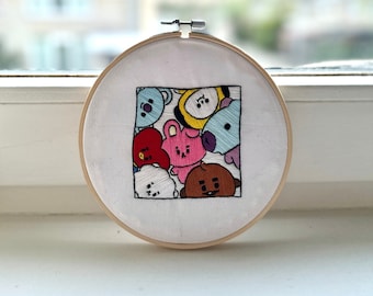 May include: Embroidered hoop art featuring colorful cartoon characters. The design is set within a square on white fabric, framed by a wooden embroidery hoop. The characters are in various colors, including blue, yellow, pink, and brown.