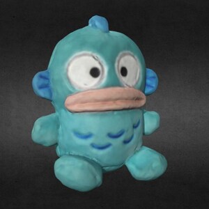 Op de afbeelding: Een turquoise cartoonwezen met grote witte ogen, een lichtroze snavel en kleine blauwe vinnen. Het pluche speelgoed heeft een afgerond lichaam en kleine armen en benen. Het wezen heeft een vin bovenop zijn hoofd en blauwe markeringen op zijn lichaam.