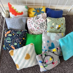 Könnte beinhalten: Eine Sammlung kleiner, quadratischer Fleece-Kissen in verschiedenen Farben und Mustern. Designs umfassen Blumen, Bienen, Dinosaurier und geometrische Formen. Die Kissen sind in einem Drahtkorb und auf einer Teppichoberfläche angeordnet.