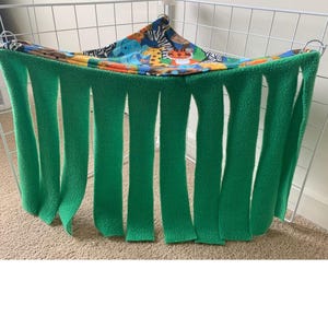 Puede incluir: Una hamaca de forro polar verde con un interior estampado de animales de colores. La hamaca tiene bordes largos con flecos y está diseñada para colgar dentro de una jaula. El estampado de animales presenta varios animales de dibujos animados.