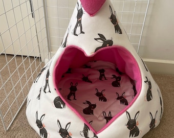 Peut inclure: Une tente pour animal de compagnie blanche avec un motif répétitif de lapins noirs portant de petits nœuds roses. L'entrée est en forme de cœur avec une doublure intérieure rose vif et un grand cœur en peluche rose au sommet.