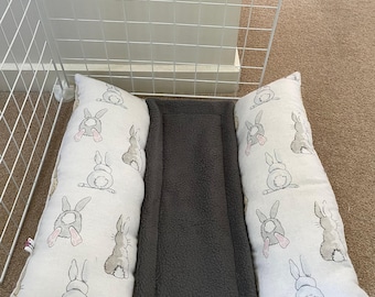May include: A pet bed with a gray fleece center and two white pillows with a bunny print. The pillows are rectangular and have a repeating pattern of bunny illustrations. The bed is designed to fit inside a wire cage.