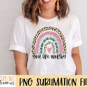 May include: A white t-shirt with a rainbow design featuring a pink glitter background and leopard print accents. The rainbow has the words "You are brave", "You are strong", and "You are loved" written on it in turquoise text. The text "You are worthy" is written below the rainbow in black text.