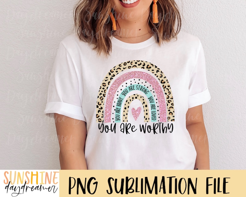 May include: A white t-shirt with a rainbow design featuring a pink glitter background and leopard print accents. The rainbow has the words "You are brave", "You are strong", and "You are loved" written on it in turquoise text. The text "You are worthy" is written below the rainbow in black text.