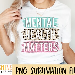 May include: White t-shirt with the text "Mental Health Matters" in black lettering. The text is layered with a light blue brushstroke, a leopard print pattern, and a pink glitter brushstroke.