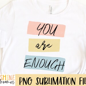May include: A white t-shirt with the text "YOU are ENOUGH" in black lettering on a colorful background. The text is in a handwritten style.