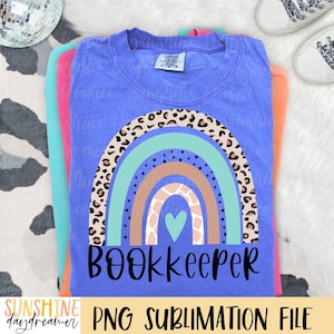 May include: A blue short-sleeved t-shirt featuring a graphic of a multi-colored rainbow with leopard print, solid teal, solid tan, and giraffe print arches. A teal heart is centered below the rainbow, and the word BOOKKEEPER is printed in black.