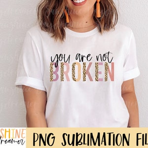 May include: White t-shirt with the text "you are not BROKEN" in black lettering. The text is layered with a pink, orange, and brown leopard print pattern.