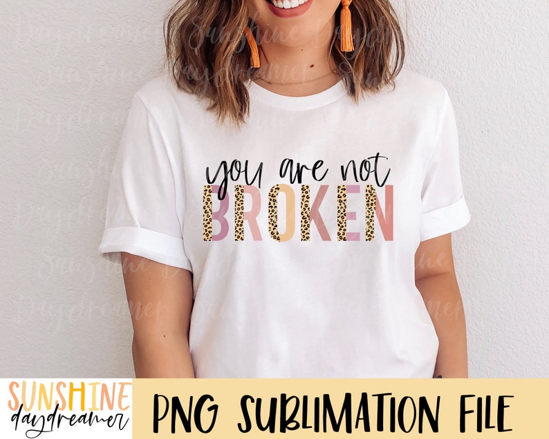 May include: White t-shirt with the text "you are not BROKEN" in black lettering. The text is layered with a pink, orange, and brown leopard print pattern.