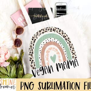 May include: A white tote bag featuring a rainbow design with a green, brown, and pink colour scheme. The rainbow is decorated with a leopard print pattern and a heart. The text "Vegan Mama" is written in black below the rainbow.