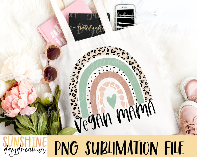 May include: A white tote bag featuring a rainbow design with a green, brown, and pink colour scheme. The rainbow is decorated with a leopard print pattern and a heart. The text "Vegan Mama" is written in black below the rainbow.