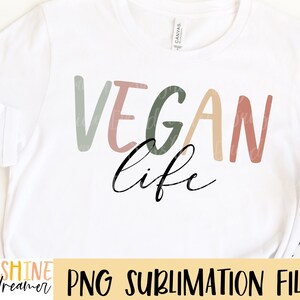 May include: A white t-shirt featuring the text "VEGAN life" in a colourful, hand-drawn style. The text is in shades of light green, pink, peach, and brown. The words "VEGAN" and "life" are separated by a line break. The text is centred on the shirt.