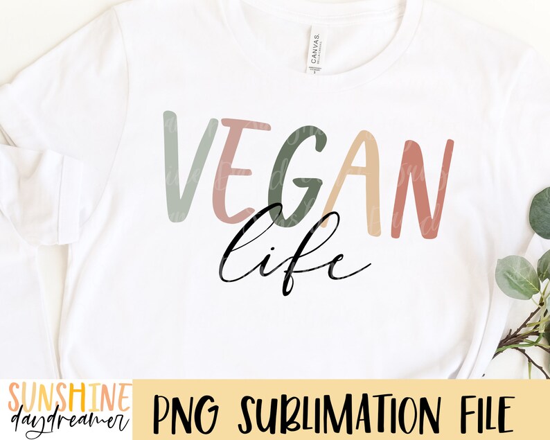 May include: A white t-shirt featuring the text "VEGAN life" in a colourful, hand-drawn style. The text is in shades of light green, pink, peach, and brown. The words "VEGAN" and "life" are separated by a line break. The text is centred on the shirt.