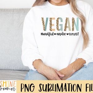 May include: White sweatshirt with the word "VEGAN" in blue and brown leopard print letters. The text "mindful aware coexist" is written below the word "VEGAN" in black.