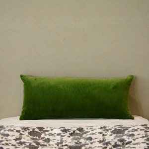 May include: A rectangular, bright green velvet cushion rests on a bed with a floral patterned duvet cover. The cushion is centered and takes up most of the frame, with a neutral wall in the background.
