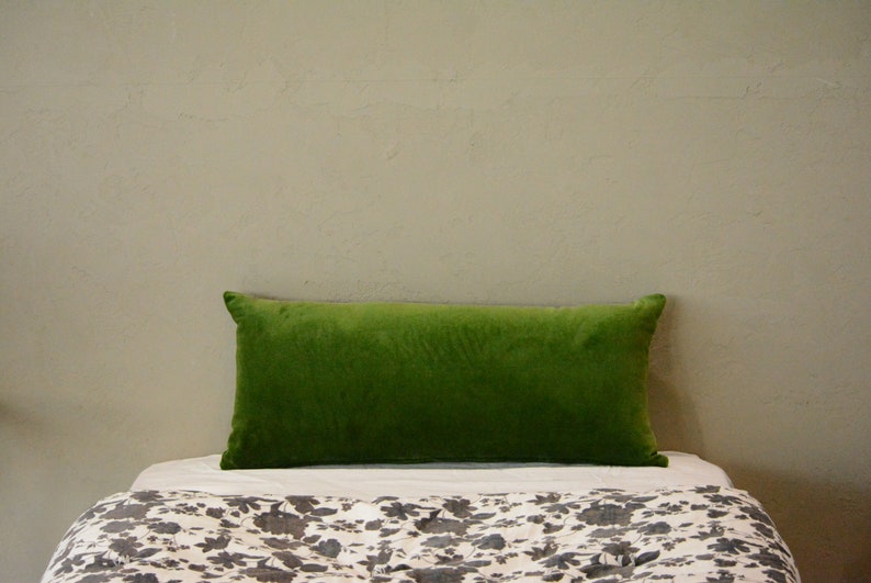 May include: A rectangular, bright green velvet cushion rests on a bed with a floral patterned duvet cover. The cushion is centered and takes up most of the frame, with a neutral wall in the background.