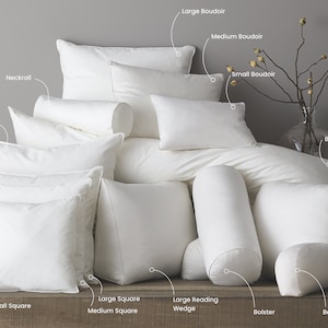 May include: An assortment of white pillows in various shapes and sizes, including square, rectangular, and cylindrical. The pillows are arranged on a wooden surface with labels identifying each type.