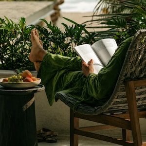 May include: A pair of loose-fitting olive green trousers and a matching long-sleeved top, worn while reading an open book. A white bowl containing green grapes and orange peaches sits on a dark cylindrical wooden side table nearby.