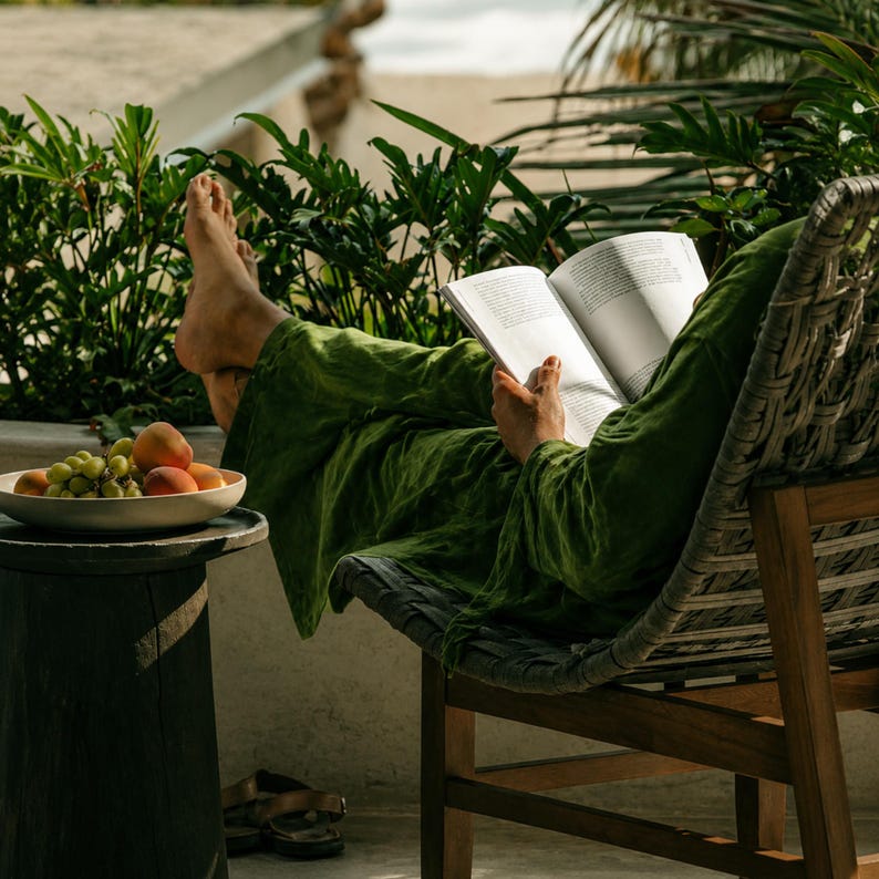 May include: A pair of loose-fitting olive green trousers and a matching long-sleeved top, worn while reading an open book. A white bowl containing green grapes and orange peaches sits on a dark cylindrical wooden side table nearby.