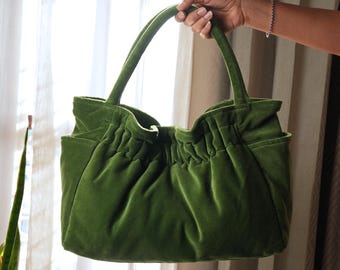 May include: A green velvet tote bag with two handles and a gathered top. The bag has a soft, plush texture and a pocket on the side. The bag is held up by a person's hand.