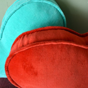 Pode incluir: Close-up de duas almofadas em forma de cora&ccedil;&atilde;o. Uma &eacute; de uma cor verde-azulado vibrante, e a outra &eacute; de um vermelho-alaranjado profundo. Ambas as almofadas t&ecirc;m uma textura macia e aveludada e s&atilde;o bordadas com uma guarni&ccedil;&atilde;o a condizer. As almofadas s&atilde;o provavelmente decorativas.