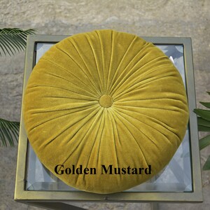 May include: A round, golden mustard-colored velvet cushion with a buttoned center and radial stitching. The cushion is placed on a glass-topped table with a gold-colored metal frame. The text "Golden Mustard" is displayed on the cushion.