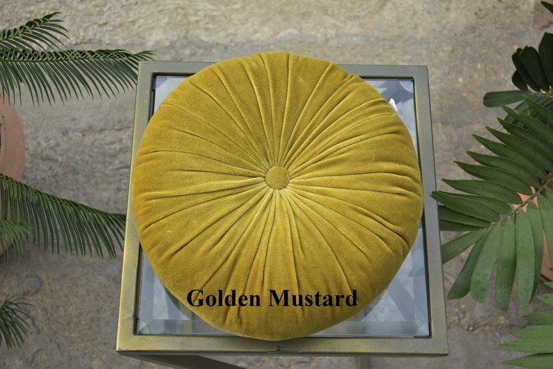 May include: A round, golden mustard-colored velvet cushion with a buttoned center and radial stitching. The cushion is placed on a glass-topped table with a gold-colored metal frame. The text "Golden Mustard" is displayed on the cushion.