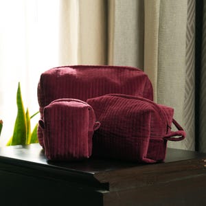 May include: Three burgundy corduroy cosmetic bags of varying sizes are arranged on a dark surface. The bags have a textured, ribbed appearance. The largest bag is rectangular, while the others are more square-shaped.
