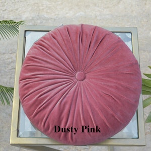 May include: A round, dusty pink velvet cushion with a button center. The cushion has a radial design and is placed on a glass-topped table with a gold frame. The text "Dusty Pink" is visible on the cushion.